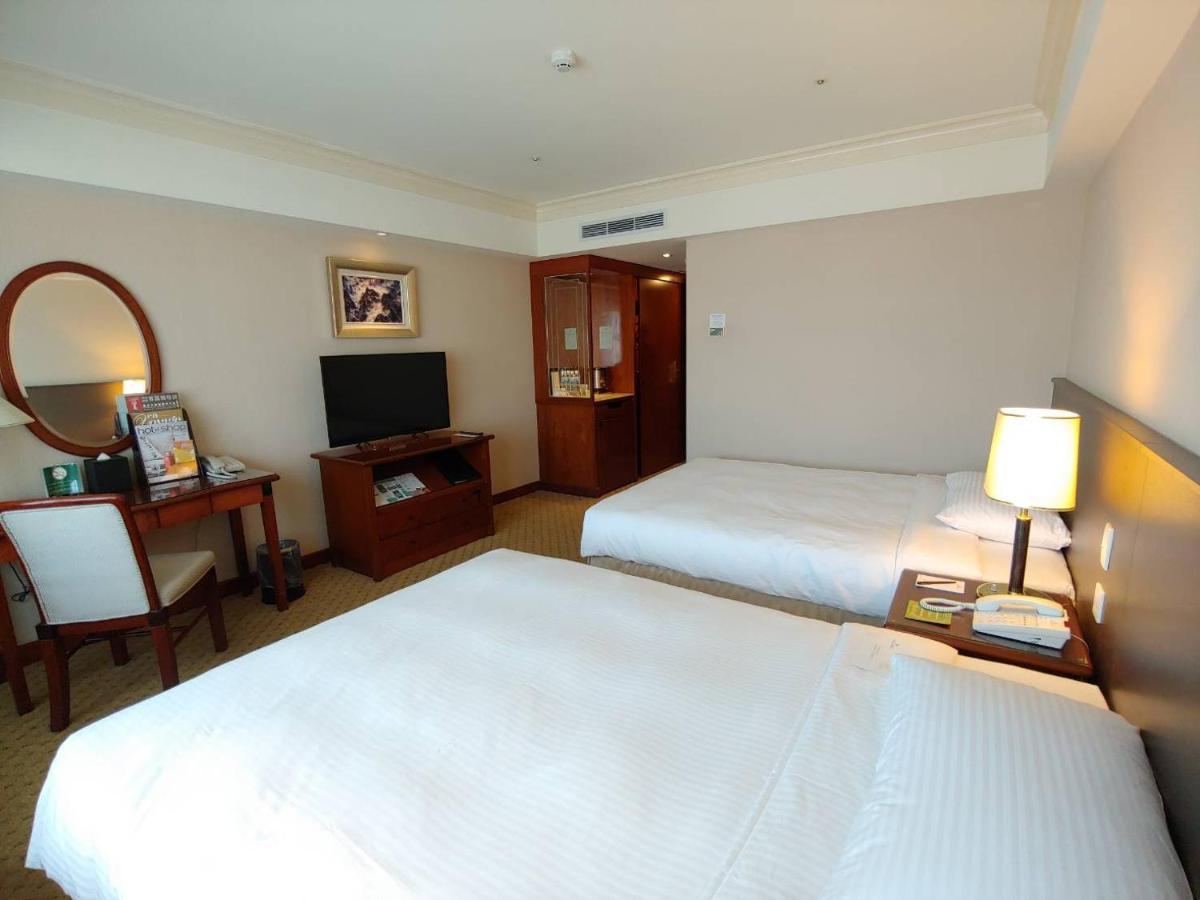 Evergreen Laurel Hotel - Taichung Exteriör bild The photo shows a hotel room with two beds, neatly made with white linens. There is a desk and chair in one corner, accompanied by a flat-screen TV mounted on the wall. A large mirror and a wooden cabinet are also visible. The room is illuminated by 